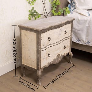 May include: A two-drawer chest with a distressed white and grey paint finish. The chest has a curved front and ornate detailing on the bottom. It measures 82 cm tall, 92 cm wide, and 40 cm deep.