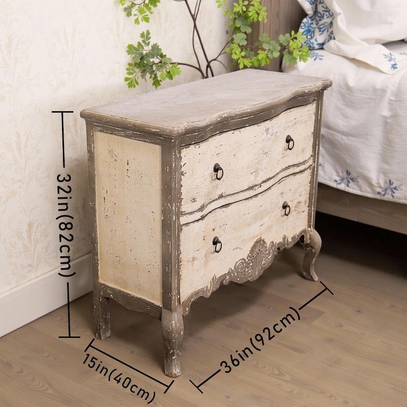May include: A two-drawer chest with a distressed white and grey paint finish. The chest has a curved front and ornate detailing on the bottom. It measures 82 cm tall, 92 cm wide, and 40 cm deep.