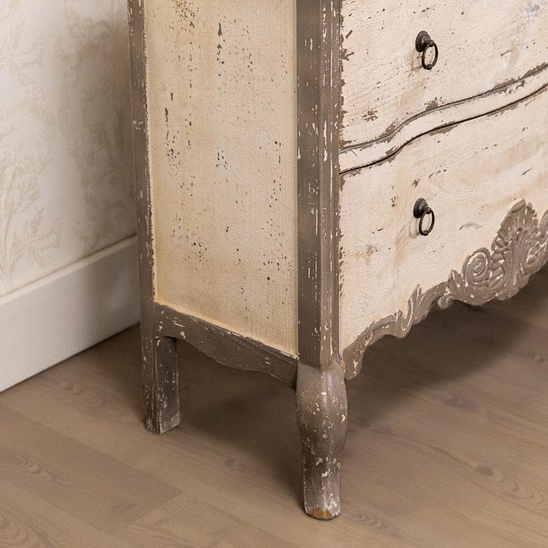 May include: A white painted wooden chest of drawers with ornate details and grey trim. The chest has two drawers with black metal handles. The chest is standing on a wooden floor.