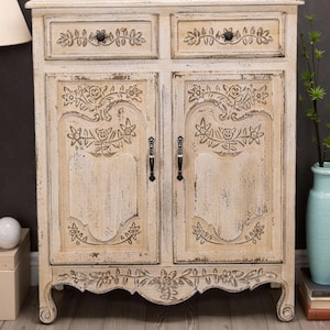 May include: A white distressed wooden cabinet with two drawers and two doors. The cabinet has carved floral details and black metal handles. The cabinet is sitting on a light wood floor.