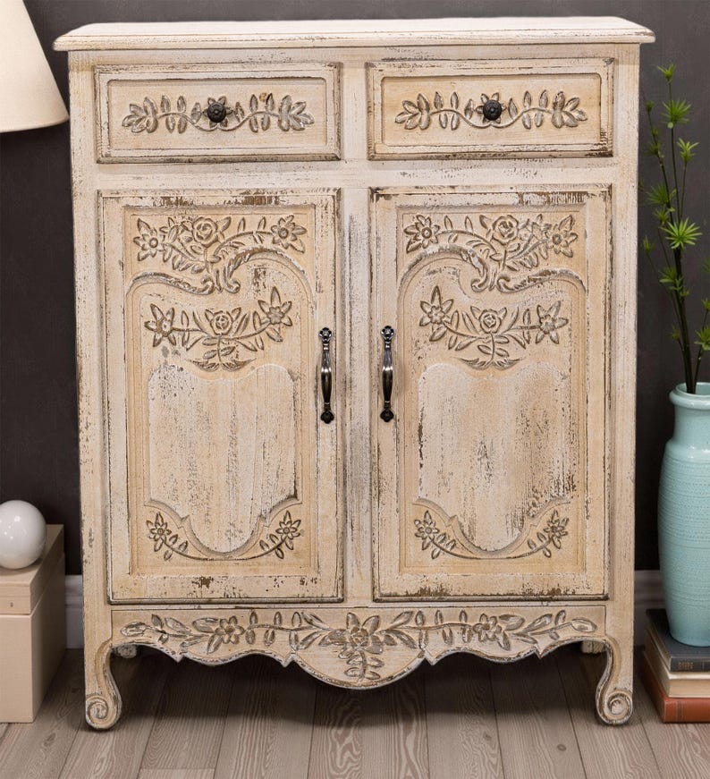 May include: A white distressed wooden cabinet with two drawers and two doors. The cabinet has carved floral details and black metal handles. The cabinet is sitting on a light wood floor.