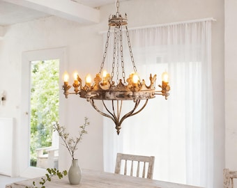 May include: A vintage-style chandelier with a crown design, hanging above a wooden table. The chandelier has multiple light bulbs and a distressed, off-white finish. The room is bright with natural light, and a vase with greenery sits on the table.