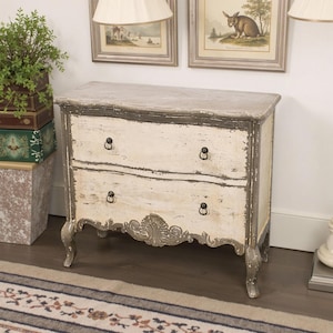 Può includere: Un comò a due cassetti con una finitura in vernice bianca e grigia invecchiata. Il comò ha un fronte curvo e intagli decorativi sul cassetto inferiore. Il comò è appoggiato su un tappeto a righe.