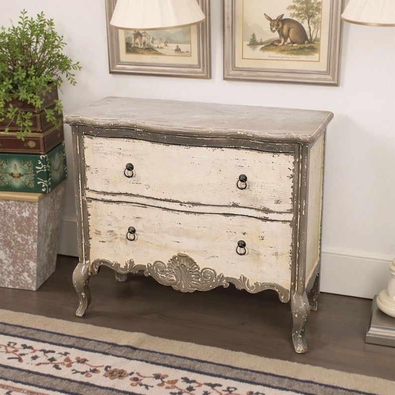 May include: A two-drawer chest with a distressed white and grey paint finish. The chest has a curved front and ornate carvings on the bottom drawer. The chest is sitting on a rug with a striped pattern.