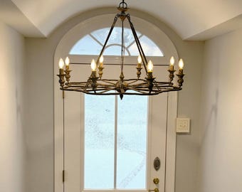 May include: A large, circular, wrought iron chandelier with 10 candle-style light bulbs. The chandelier is hanging from a white ceiling in a hallway with a white door and a window.