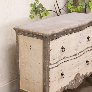 May include: A distressed white and grey wooden chest of drawers with ornate carvings and black metal drawer pulls.