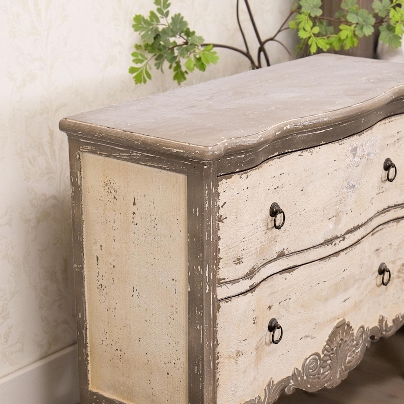 May include: A distressed white and grey wooden chest of drawers with ornate carvings and black metal drawer pulls.