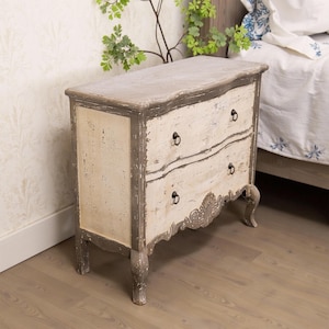 May include: A two-drawer chest of drawers with a distressed white and grey paint finish. The chest has a curved top and ornate carvings on the bottom drawer. It has black metal drawer pulls.