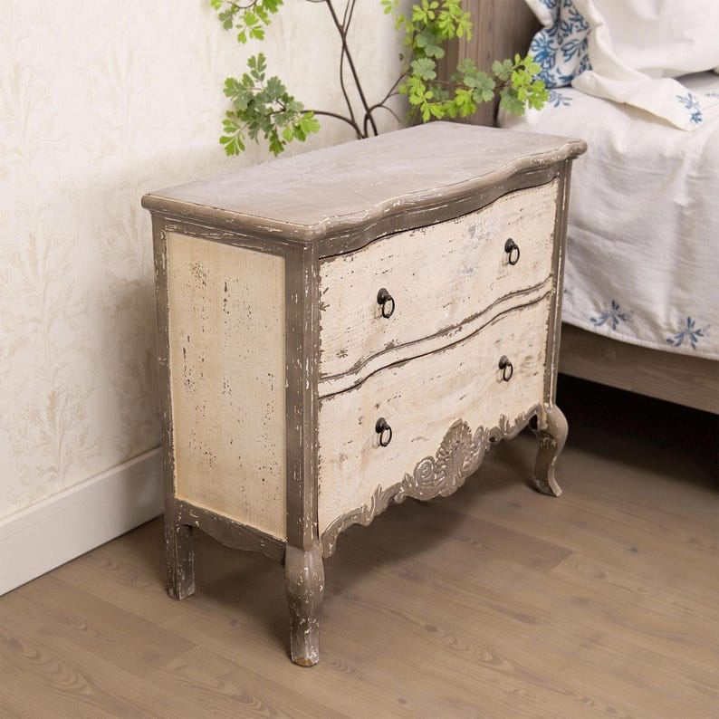 May include: A two-drawer chest of drawers with a distressed white and grey paint finish. The chest has a curved top and ornate carvings on the bottom drawer. It has black metal drawer pulls.