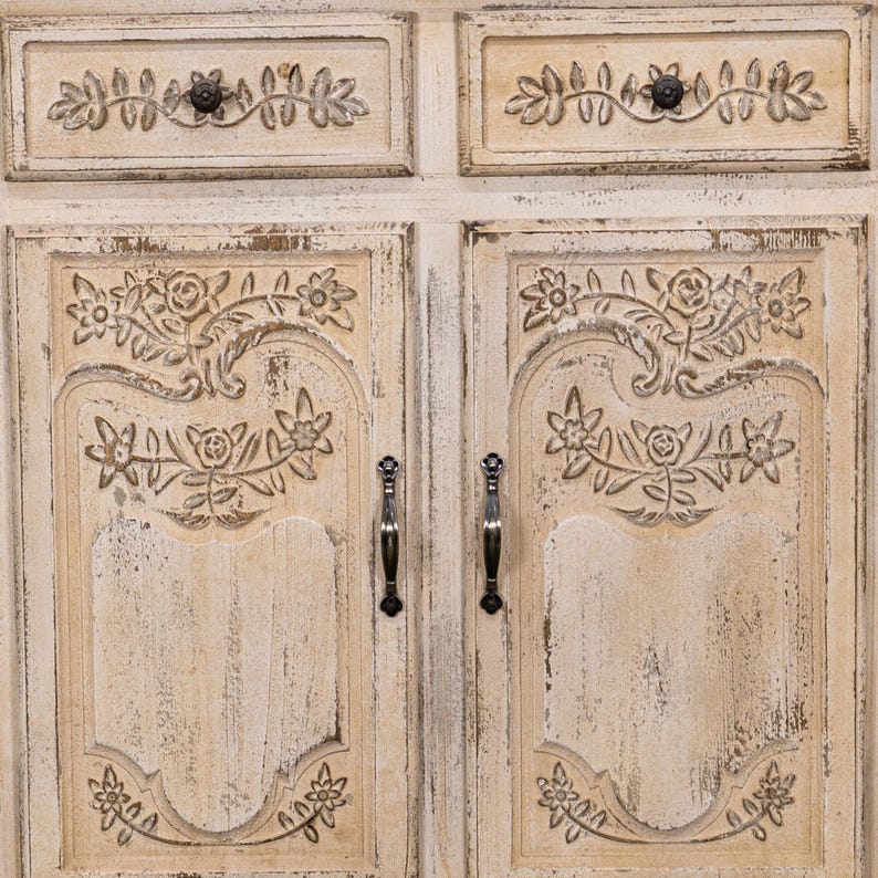 May include: A white distressed wooden cabinet with two doors and two drawers. The doors and drawers have carved floral designs and black metal handles.