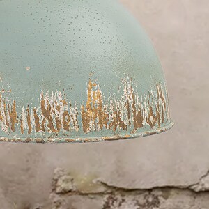 May include: Close-up of a distressed, light blue metal pendant light. The shade has a domed shape with a textured surface and a worn, gold-colored trim, giving it a vintage appearance. The light fixture is likely for home decor.