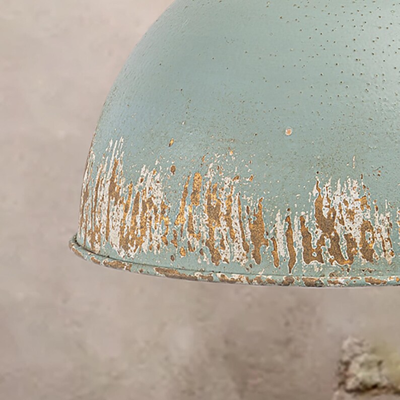 May include: Close-up of a distressed, light blue metal pendant light. The lamp has a dome shape with a weathered, textured finish. The edges show areas of exposed gold-colored metal, adding to the vintage aesthetic. The lamp has small perforations across the surface.