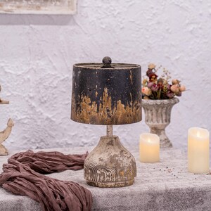 May include: A distressed black and gold lamp with a wooden base. The lamp is on a table with a brown fabric and two white candles.