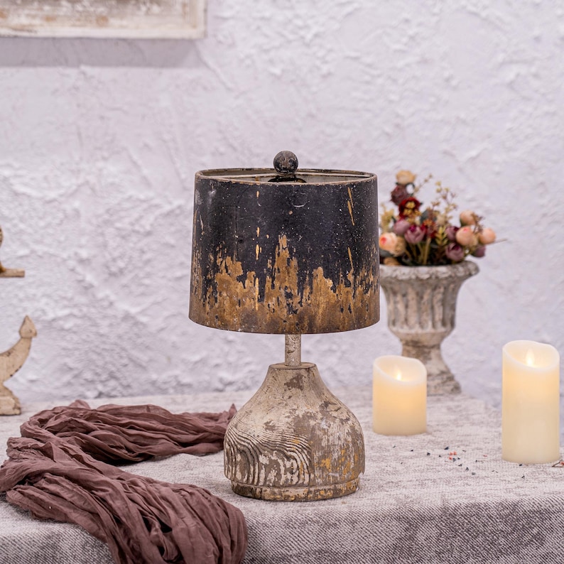 May include: A distressed black and gold lamp with a wooden base. The lamp is on a table with a brown fabric and two white candles.