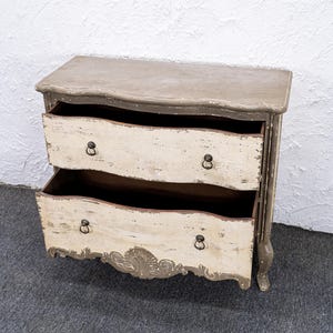 May include: A two-drawer chest of drawers with a distressed white and grey paint finish. The chest has a curved front and ornate carvings on the bottom.