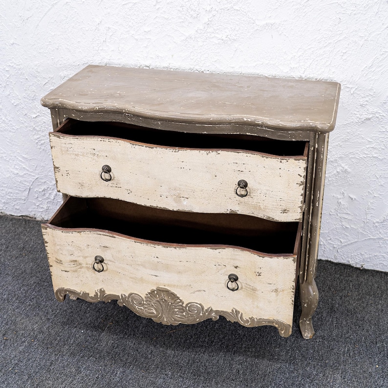 May include: A two-drawer chest of drawers with a distressed white and grey paint finish. The chest has a curved front and ornate carvings on the bottom.