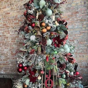 May include: A decorated Christmas tree with red, green, and gold ornaments, including baubles, bells, and poinsettias. The tree is adorned with a wooden sign that reads "JOY". The tree is set against a brick wall.