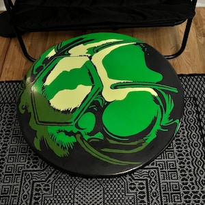 May include: A round, black and green decorative object with a stylized graphic design. The design features a green skull-like image with light green and white accents. The object is sitting on a black and white patterned rug.