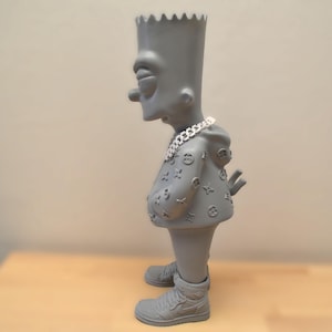 Peut inclure: Une figurine grise d'un personnage de dessin animé portant une veste à capuche avec un motif et un collier en chaîne argentée. La figurine est debout sur une surface en bois.