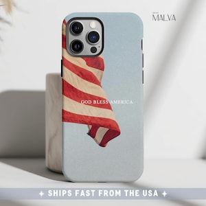 May include: A light blue smartphone case featuring a graphic of a waving American flag and the text GOD BLESS AMERICA printed in white. The case has a cutout for a triple-lens camera system on the upper left side and a smooth matte finish.