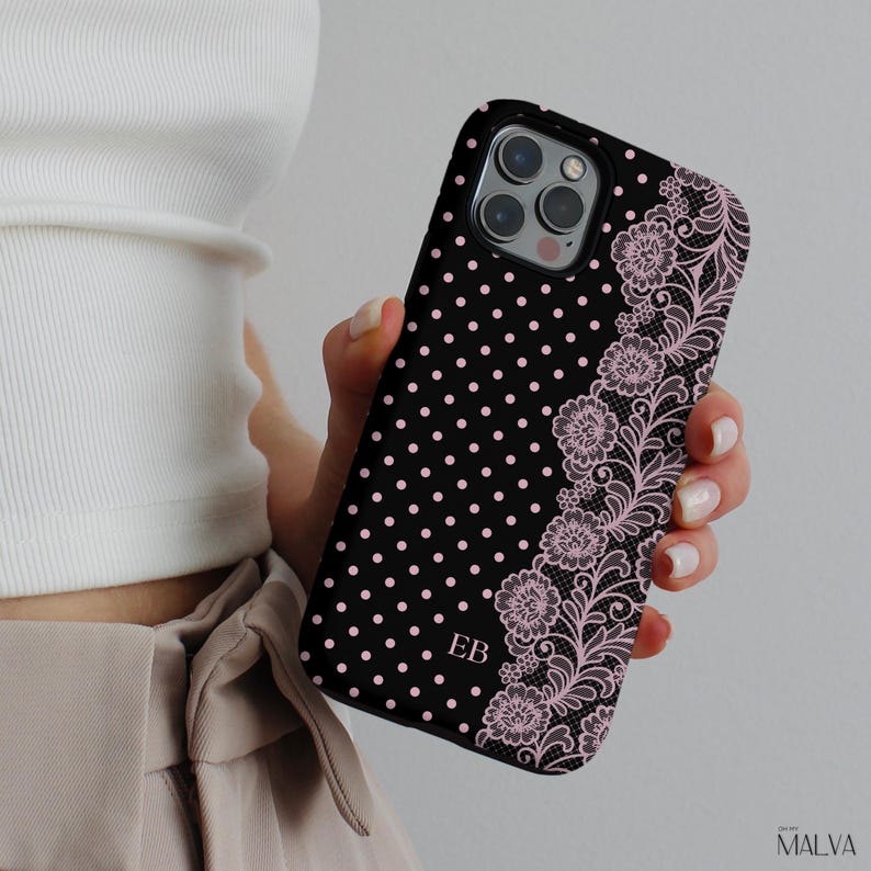 May include: A black smartphone case featuring a pattern of small pink polka dots across the left side and a detailed pink floral lace design along the right edge. The letters EB are printed in a simple font in the lower center of the case.