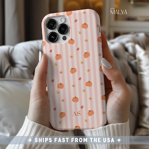 May include: A phone case featuring a pattern of small orange pumpkins and tiny yellow leaves over a background of vertical light pink and white stripes. The lower center of the case displays the capitalized letters AS in a simple serif font.