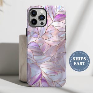 May include: A phone case with a stained glass leaf design in shades of purple, pink, and white, outlined with gold. The case has a glossy finish and a multi-camera lens cutout. The text "SHIPS FAST" is visible.