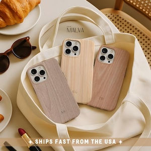 May include: Three rectangular smartphone cases with a wood grain texture and engraved initials AS, KG, and TD. The cases are arranged inside a cream-colored canvas tote bag. Each case features a circular camera cutout with three lenses.