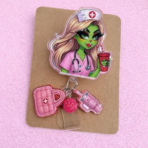 May include: A badge reel featuring a cartoon nurse with green skin, blonde hair, and a pink uniform. The reel includes a pink mug, a pink glitter ball, and a pink syringe charm. The nurse is holding a red cup with a green creature design.