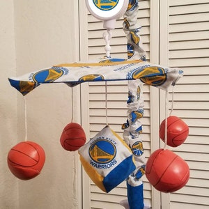 May include: A baby mobile with a basketball theme. The mobile features a round, white, plastic base with a Golden State Warriors logo. The mobile has five hanging elements: four red basketballs and a yellow and blue fabric square with the Golden State Warriors logo.