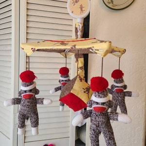 May include: A baby mobile with a yellow and brown fabric canopy and four knitted monkey toys hanging from it. The monkeys are wearing red pom-poms on their heads and have white faces. The mobile is hanging from a white metal frame.