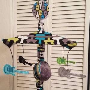 May include: A baby mobile with a black, white, green and blue patterned canopy. The mobile has four hanging toys: a grey and white striped ball, a black electric guitar, a green electric guitar and a silver electric guitar.