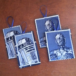 May include: Four handmade fabric ornaments featuring characters from Star Wars. Two ornaments feature R2-D2, a droid with a blue and white design. Two ornaments feature C-3PO, a droid with a gold design.