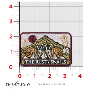 May include: An embroidered patch with a white background and a burgundy border. The patch features two snails with brown shells and green bodies, climbing a green mountain with a red sun in the background. The text "THE RUSTY SNAILS" is written in brown letters below the snails.