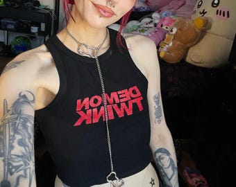 May include: A black tank top with the red text "DEMON INK" is worn with a silver chain necklace. The necklace has a bone-shaped pendant. The person has red and black hair and multiple piercings.