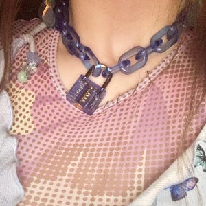May include: A close-up of a blue chain necklace with a clear padlock clasp. The necklace is worn over a pink dotted top and a light blue top with butterfly graphics.