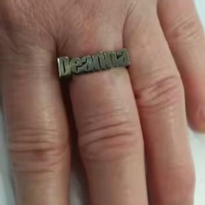 Op de afbeelding: Een zilverkleurige ring met de naam "Deanna" in blokletters. De ring wordt aan een vinger gedragen en toont het gepersonaliseerde sieraad. Het ontwerp van de ring is eenvoudig en elegant, geschikt voor dagelijks gebruik.