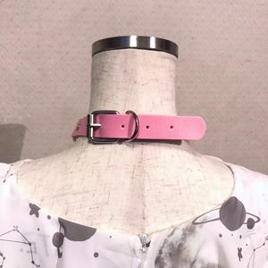 May include: A pink faux leather choker with a silver buckle and two silver rings. The choker is on a white mannequin with a white and gray patterned shirt.