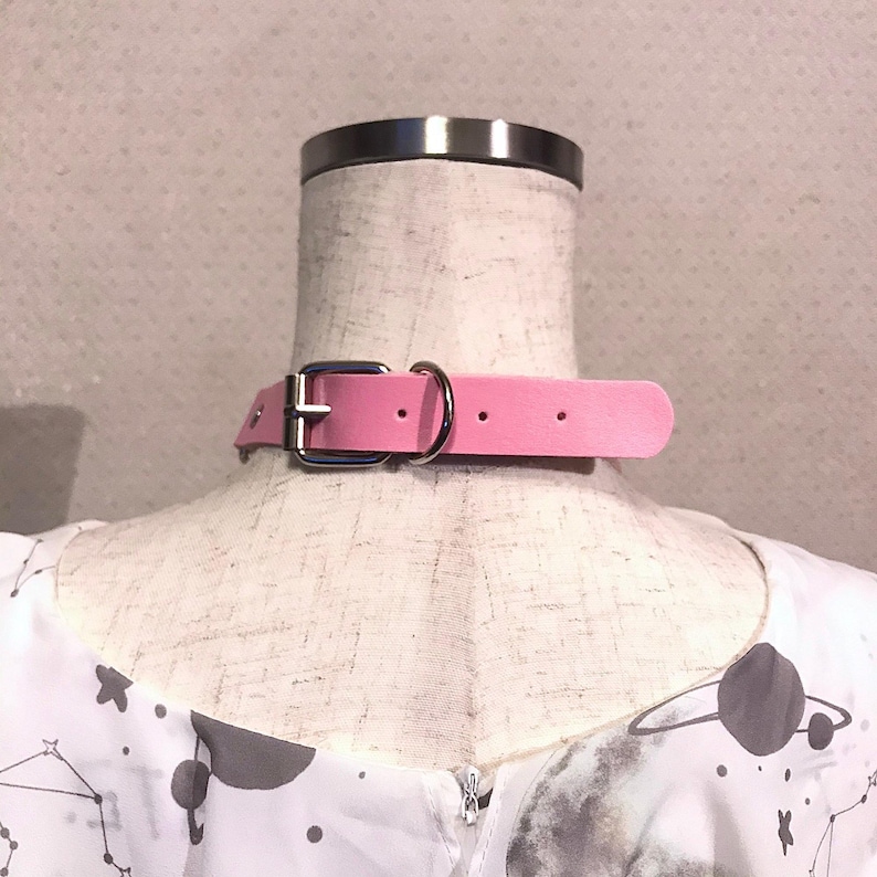 May include: A pink faux leather choker with a silver buckle and two silver rings. The choker is on a white mannequin with a white and gray patterned shirt.
