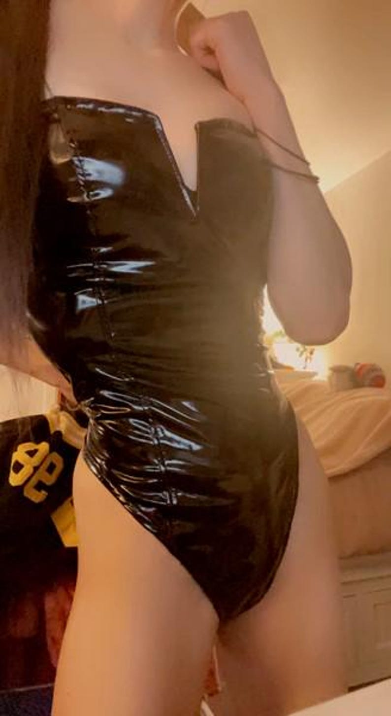 May include: A shiny, black bodysuit with a deep V-neckline. The one-piece garment has a glossy, wet-look finish. The strapless design is form-fitting and appears to be made of a synthetic material. The bodysuit is a bold fashion statement.