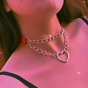 May include: A silver chain choker necklace with a large heart pendant. The chain is double layered and has a red leather strap.