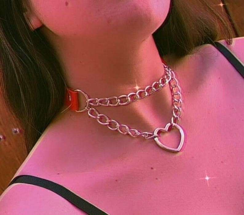 May include: A silver chain choker necklace with a large heart pendant. The chain is double layered and has a red leather strap.