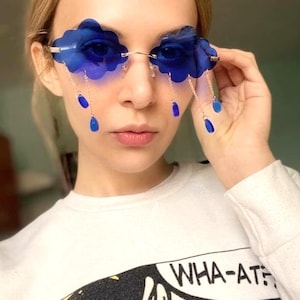 May include: Blue cloud-shaped sunglasses with dangling blue raindrop charms. The sunglasses have a gold frame and are worn by a person with blonde hair. The person is wearing a white shirt with a comic book graphic that says "WHA-AT?"