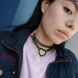 May include: A black choker necklace with a heart pendant. The necklace is made of two chains, one thicker than the other. The heart pendant is open and has a smooth finish.