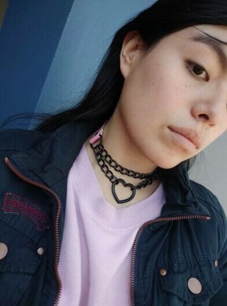 May include: A black choker necklace with a heart pendant. The necklace is made of two chains, one thicker than the other. The heart pendant is open and has a smooth finish.