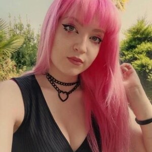 May include: A woman with bright pink hair wearing a black sleeveless top with a deep V-neckline. She is wearing a black choker necklace with a heart pendant.