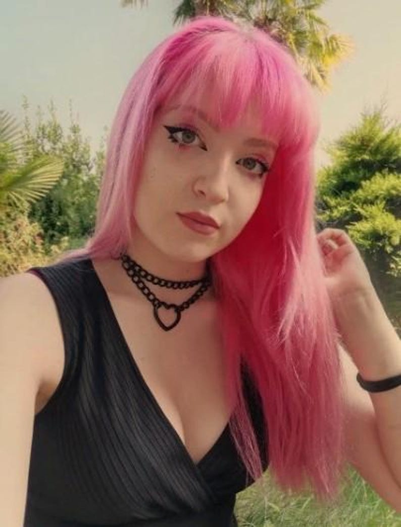 May include: A woman with bright pink hair wearing a black sleeveless top with a deep V-neckline. She is wearing a black choker necklace with a heart pendant.