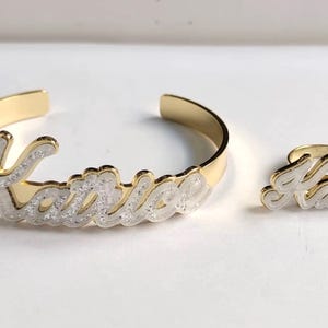Puede incluir: Conjunto de pulsera y anillo de oro con el nombre "Katrice" escrito en purpurina blanca.
