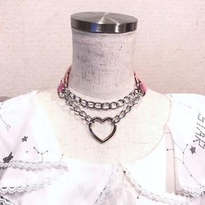May include: A silver chain choker necklace with a large heart charm. The chain is layered and has a pink leather strap.