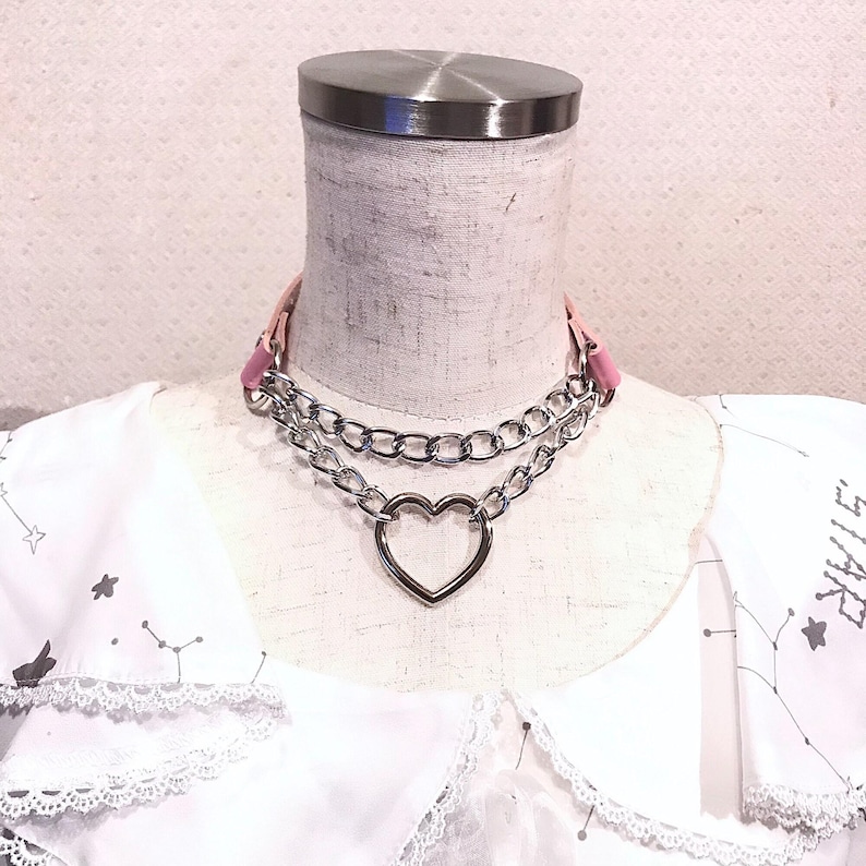 May include: A silver chain choker necklace with a large heart charm. The chain is layered and has a pink leather strap.